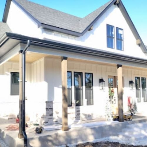 A two-story white house with a gabled roof and a front porch supported by wooden pillars and black gutters by gutter contractors in San Antonio, TX